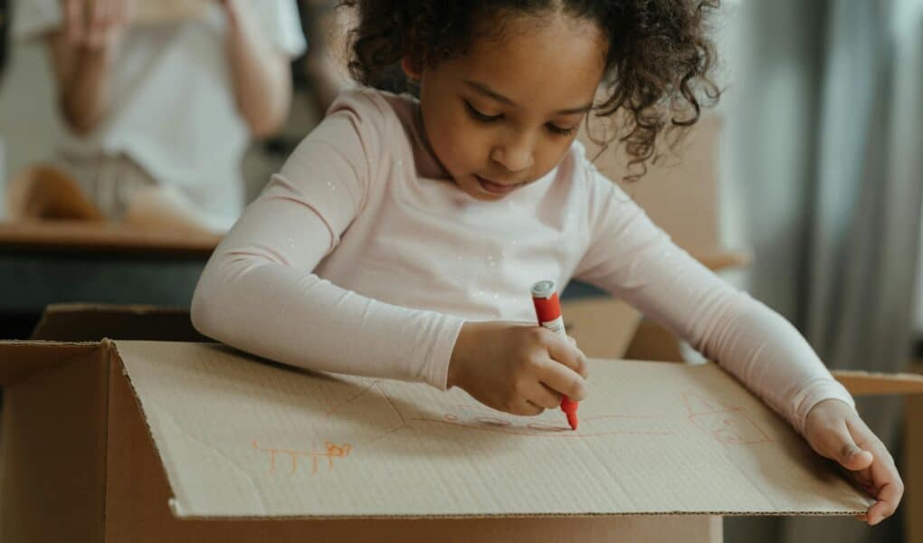 A young girl with curly hair, wearing a light pink long-sleeved shirt, sits inside a large cardboard box and uses a red felt-tip pen to draw on the box’s flap. Her expression is focused, and another person is blurred in the background.