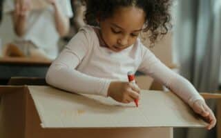 A young girl with curly hair, wearing a light pink long-sleeved shirt, sits inside a large cardboard box and uses a red felt-tip pen to draw on the box’s flap. Her expression is focused, and another person is blurred in the background.