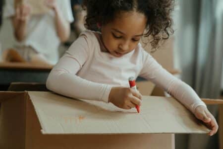 A young girl with curly hair, wearing a light pink long-sleeved shirt, sits inside a large cardboard box and uses a red felt-tip pen to draw on the box’s flap. Her expression is focused, and another person is blurred in the background.