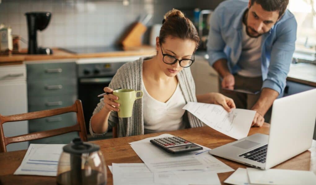 A woman with glasses sits at a kitchen table, holding a green mug and reviewing papers with a pen. A man leans over beside her, looking at the documents. A calculator, laptop, coffee pot, and papers are spread out on the wooden table.