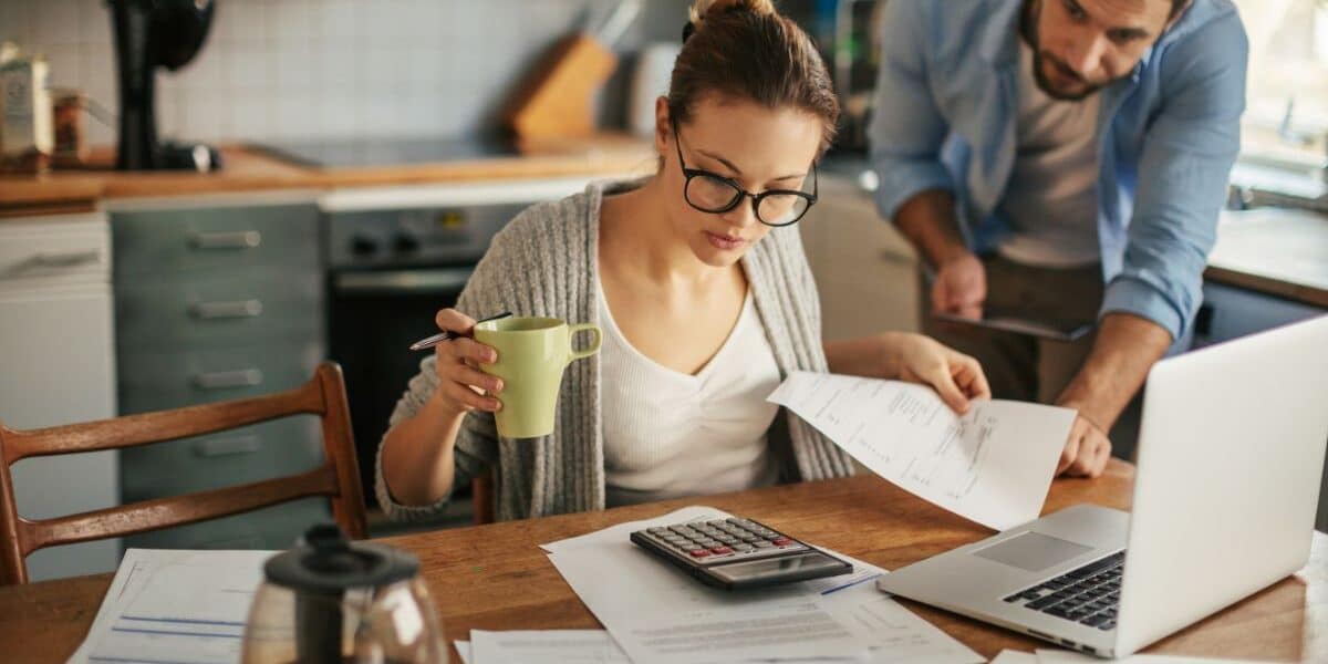 A woman with glasses sits at a kitchen table, holding a green mug and reviewing papers with a pen. A man leans over beside her, looking at the documents. A calculator, laptop, coffee pot, and papers are spread out on the wooden table.