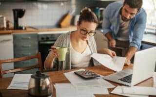 A woman with glasses sits at a kitchen table, holding a green mug and reviewing papers with a pen. A man leans over beside her, looking at the documents. A calculator, laptop, coffee pot, and papers are spread out on the wooden table.