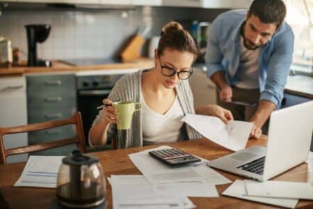 A woman with glasses sits at a kitchen table, holding a green mug and reviewing papers with a pen. A man leans over beside her, looking at the documents. A calculator, laptop, coffee pot, and papers are spread out on the wooden table.
