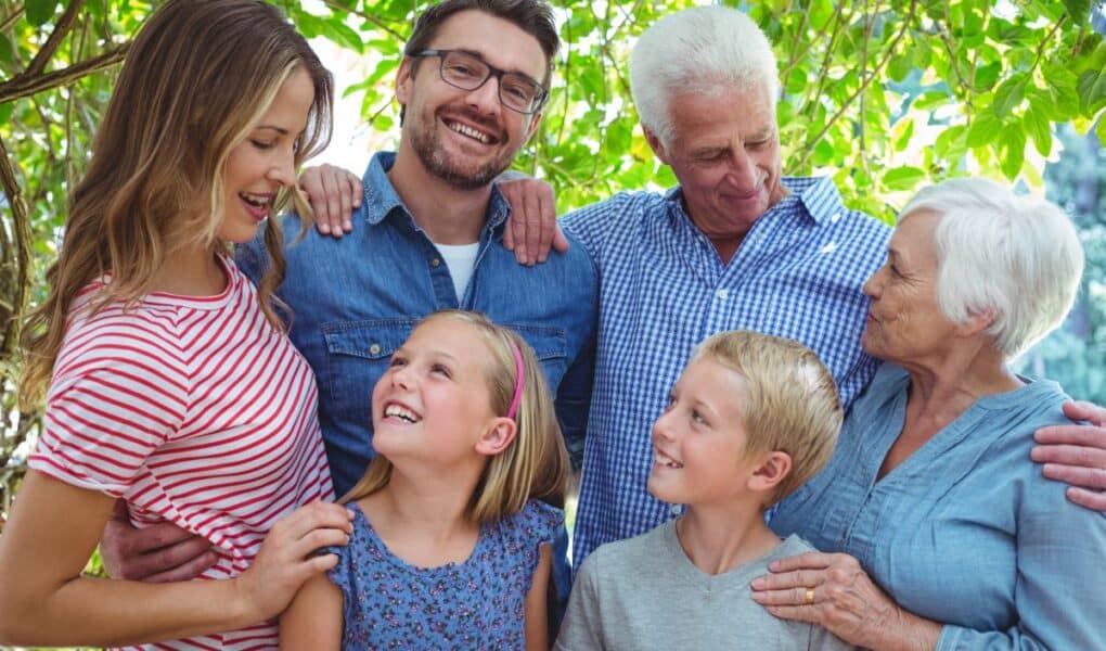 Three generations of a smiling family stand close together outdoors. Two older adults, two younger adults, and two children are surrounded by green leaves. They look at each other with joy, expressing warmth, happiness, and a sense of togetherness.