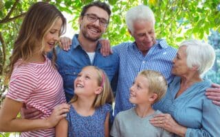 Three generations of a smiling family stand close together outdoors. Two older adults, two younger adults, and two children are surrounded by green leaves. They look at each other with joy, expressing warmth, happiness, and a sense of togetherness.