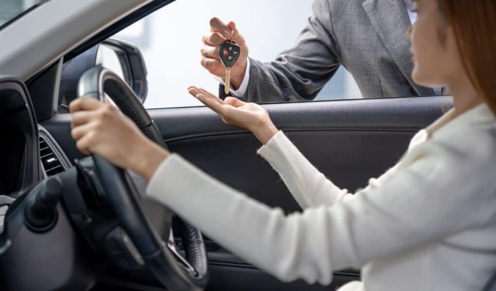A person sitting in the driver's seat of a car reaches out as another person, standing outside and wearing a suit, hands them a car key. The focus is on the exchange, with the car interior and steering wheel visible.