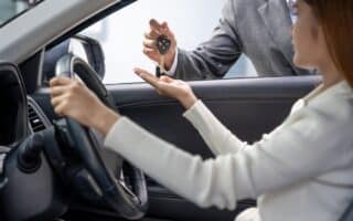 A person sitting in the driver's seat of a car reaches out as another person, standing outside and wearing a suit, hands them a car key. The focus is on the exchange, with the car interior and steering wheel visible.