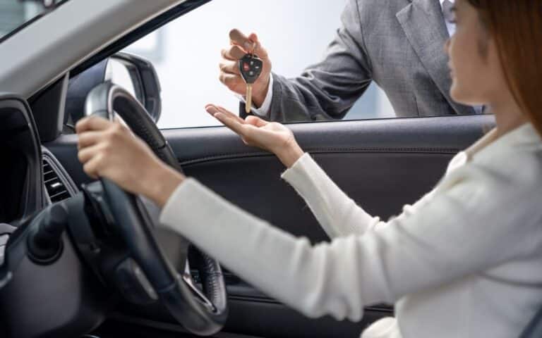 A person sitting in the driver's seat of a car reaches out as another person, standing outside and wearing a suit, hands them a car key. The focus is on the exchange, with the car interior and steering wheel visible.