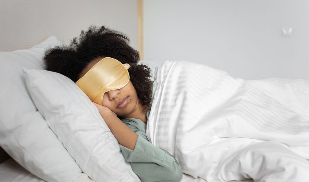 A person with curly hair is lying on their side in bed, wearing a gold sleep mask and light green pyjamas. They are covered with a white duvet and resting their head on a white pillow, appearing peaceful and relaxed in a softly lit bedroom.