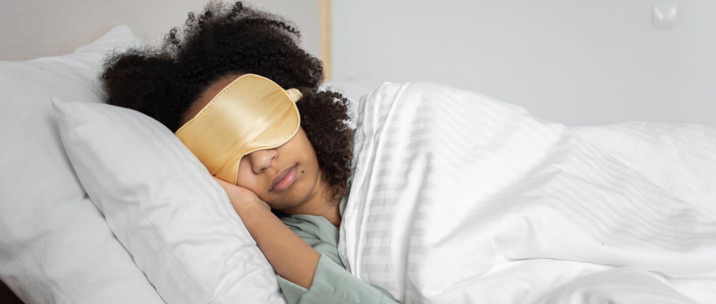 A person with curly hair is lying on their side in bed, wearing a gold sleep mask and light green pyjamas. They are covered with a white duvet and resting their head on a white pillow, appearing peaceful and relaxed in a softly lit bedroom.