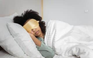 A person with curly hair is lying on their side in bed, wearing a gold sleep mask and light green pyjamas. They are covered with a white duvet and resting their head on a white pillow, appearing peaceful and relaxed in a softly lit bedroom.