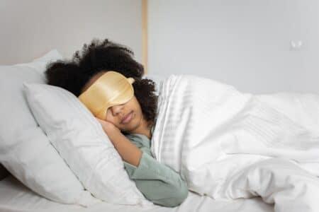 A person with curly hair is lying on their side in bed, wearing a gold sleep mask and light green pyjamas. They are covered with a white duvet and resting their head on a white pillow, appearing peaceful and relaxed in a softly lit bedroom.