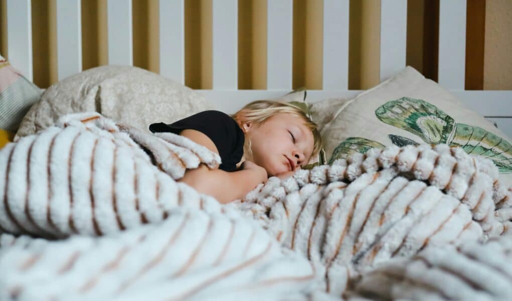 A young child with blonde hair sleeps peacefully in bed, partially covered by a fluffy, striped blanket. The child rests on soft pillows, one featuring a green leaf design. The background shows a white slatted headboard.
