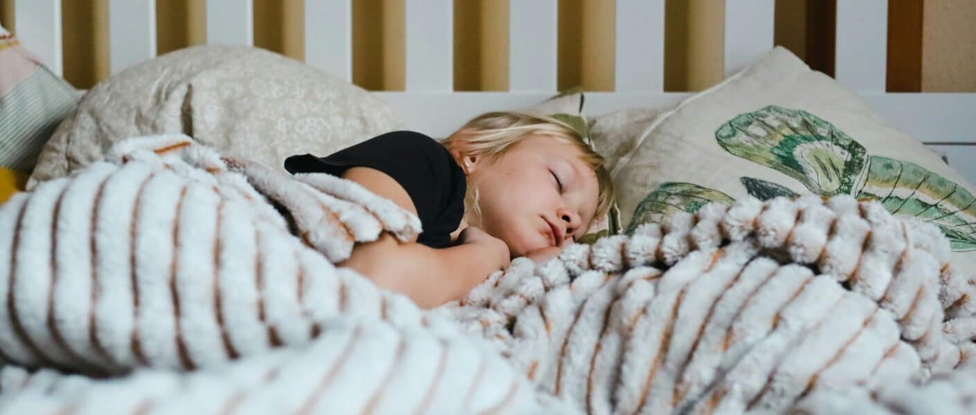 A young child with blonde hair sleeps peacefully in bed, partially covered by a fluffy, striped blanket. The child rests on soft pillows, one featuring a green leaf design. The background shows a white slatted headboard.