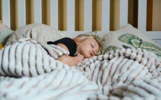 A young child with blonde hair sleeps peacefully in bed, partially covered by a fluffy, striped blanket. The child rests on soft pillows, one featuring a green leaf design. The background shows a white slatted headboard.