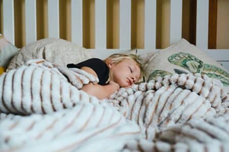 A young child with blonde hair sleeps peacefully in bed, partially covered by a fluffy, striped blanket. The child rests on soft pillows, one featuring a green leaf design. The background shows a white slatted headboard.