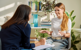 Two women sit opposite each other in a bright room with plants and bookshelves. One woman, holding a pen and clipboard, listens attentively. The other, in a white shirt and jeans, gestures while speaking. Sunlight streams through the window.