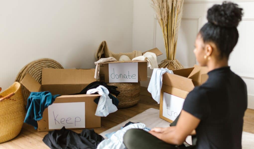 A woman sits on the floor, facing three cardboard boxes labelled “keep,” “donate,” and “rubbish.” Clothes are draped over the boxes. Decorative baskets, hats, and tall dried grasses are in the background. The setting is calm and organised.