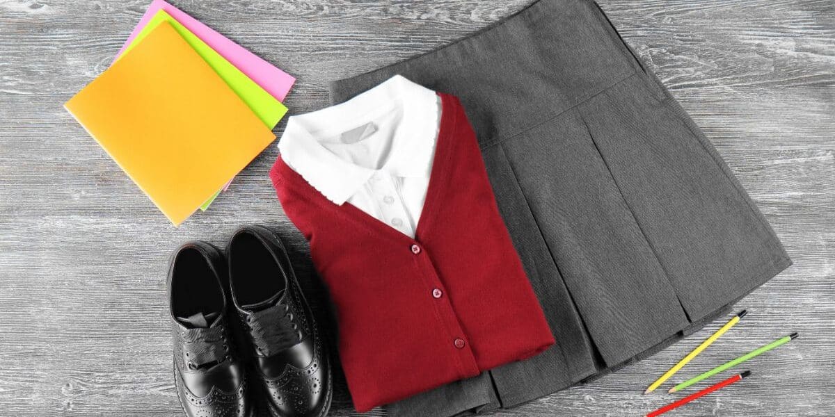 A folded grey pleated skirt, white collared shirt, and red cardigan are next to black dress shoes, three coloured pencils, and three coloured folders on a grey wood surface.