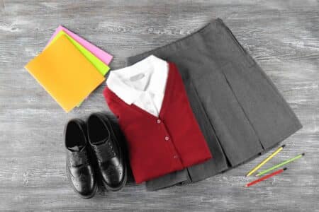 A folded grey pleated skirt, white collared shirt, and red cardigan are next to black dress shoes, three coloured pencils, and three coloured folders on a grey wood surface.