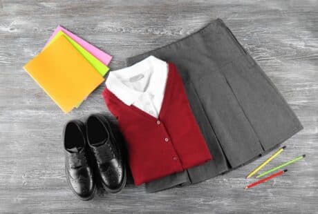 A folded grey pleated skirt, white collared shirt, and red cardigan are next to black dress shoes, three coloured pencils, and three coloured folders on a grey wood surface.