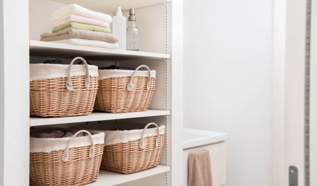 Four shelves hold wicker baskets with fabric linings, folded towels, and bottles in a white, organised cupboard. A beige towel hangs in the adjoining bathroom. The space is tidy, bright, and minimal, suggesting a laundry or linen cupboard.