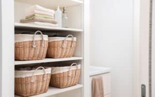 Four shelves hold wicker baskets with fabric linings, folded towels, and bottles in a white, organised cupboard. A beige towel hangs in the adjoining bathroom. The space is tidy, bright, and minimal, suggesting a laundry or linen cupboard.