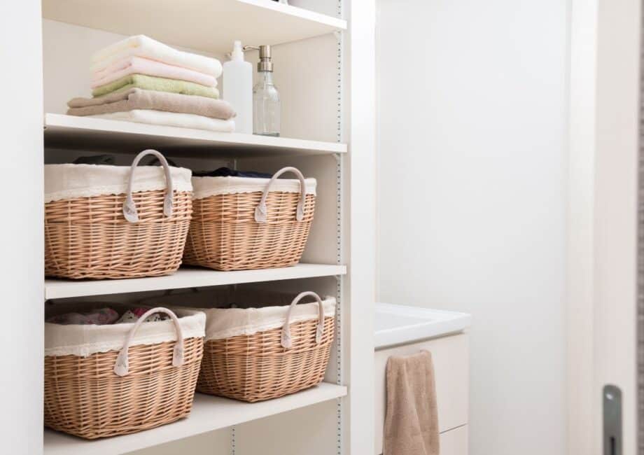 Four shelves hold wicker baskets with fabric linings, folded towels, and bottles in a white, organised cupboard. A beige towel hangs in the adjoining bathroom. The space is tidy, bright, and minimal, suggesting a laundry or linen cupboard.