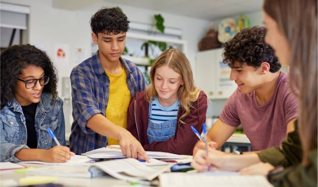 Five teenagers sit at a table covered in books and papers, working together. One boy points to a page while others look on and take notes. They are focused, collaborating in a classroom with bright lighting and educational decor.