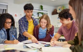 Five teenagers sit at a table covered in books and papers, working together. One boy points to a page while others look on and take notes. They are focused, collaborating in a classroom with bright lighting and educational decor.