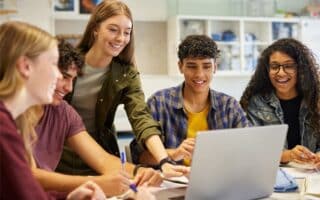 Five diverse teenagers gather around a laptop in a bright classroom, smiling and collaborating. One girl points at the screen, while others hold notebooks and pens. Science equipment is visible in the background. The atmosphere is positive and engaged.