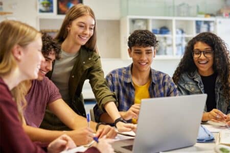 Five diverse teenagers gather around a laptop in a bright classroom, smiling and collaborating. One girl points at the screen, while others hold notebooks and pens. Science equipment is visible in the background. The atmosphere is positive and engaged.