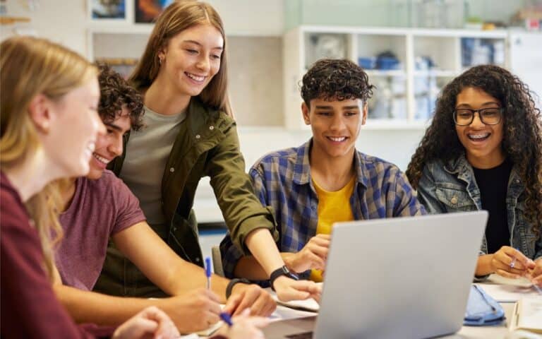 Five diverse teenagers gather around a laptop in a bright classroom, smiling and collaborating. One girl points at the screen, while others hold notebooks and pens. Science equipment is visible in the background. The atmosphere is positive and engaged.