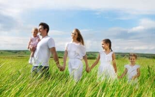 A family of five, dressed in white, walks through a green field. The father carries a toddler, whilst the mother and two girls walk hand in hand. The sky is blue with clouds, and the grass is tall, giving a feeling of warmth and togetherness.