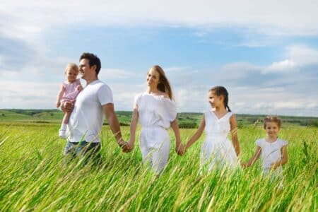 A family of five, dressed in white, walks through a green field. The father carries a toddler, whilst the mother and two girls walk hand in hand. The sky is blue with clouds, and the grass is tall, giving a feeling of warmth and togetherness.