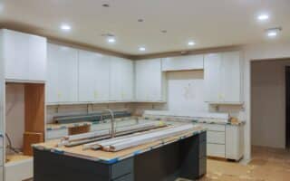 A modern kitchen under construction with white cupboards, a centre island with a sink, light fittings, and unfinished surfaces. Cardboard and tape cover worktops, and tools and wooden planks are on the island. No appliances are installed yet.