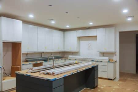 A modern kitchen under construction with white cupboards, a centre island with a sink, light fittings, and unfinished surfaces. Cardboard and tape cover worktops, and tools and wooden planks are on the island. No appliances are installed yet.