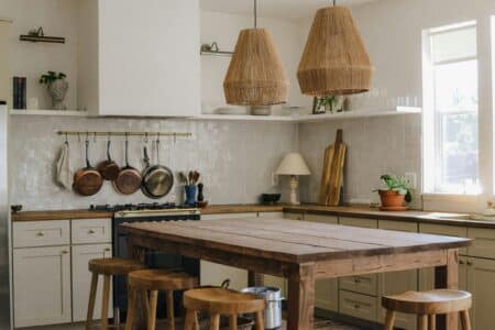 A cosy kitchen with white cupboards, wooden worktops, a rustic wooden table, and four stools. Pots and pans hang above a cooker. Two woven pendant lights hang from the ceiling. Light streams in through two windows on the right, illuminating the space.