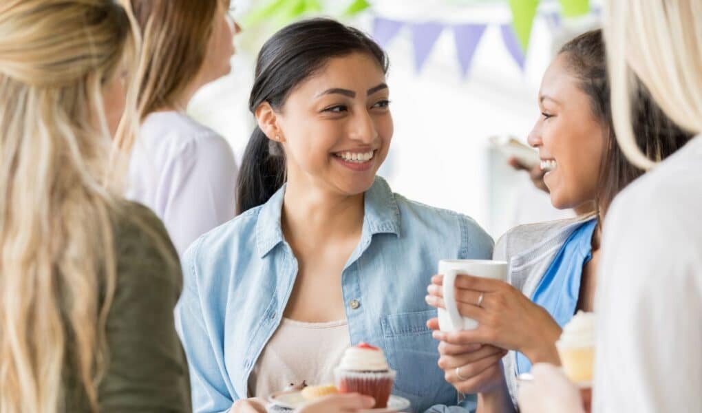 Four women stand together at a gathering, smiling and holding cupcakes and mugs. One woman in a blue shirt is looking at another woman with a ponytail, who holds a white mug. Colourful bunting is hanging in the background, creating a festive mood.
