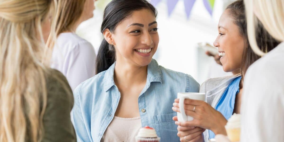 Four women stand together at a gathering, smiling and holding cupcakes and mugs. One woman in a blue shirt is looking at another woman with a ponytail, who holds a white mug. Colourful bunting is hanging in the background, creating a festive mood.
