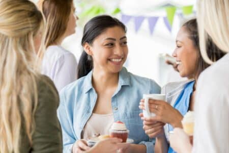 Four women stand together at a gathering, smiling and holding cupcakes and mugs. One woman in a blue shirt is looking at another woman with a ponytail, who holds a white mug. Colourful bunting is hanging in the background, creating a festive mood.