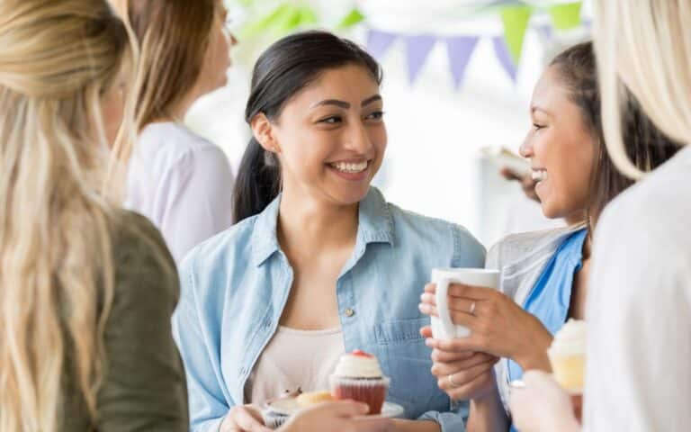 Four women stand together at a gathering, smiling and holding cupcakes and mugs. One woman in a blue shirt is looking at another woman with a ponytail, who holds a white mug. Colourful bunting is hanging in the background, creating a festive mood.