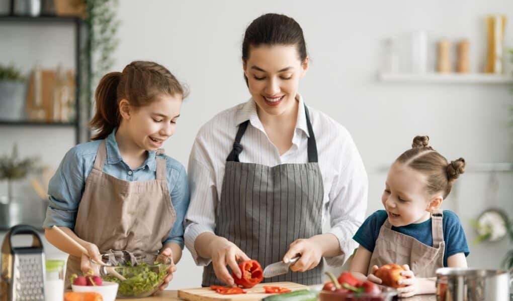 A woman and two young girls, all wearing aprons, prepare food together in a bright kitchen. The woman slices a red pepper while one girl mixes a salad and the other smiles, holding a tomato. Fresh vegetables are on the counter.