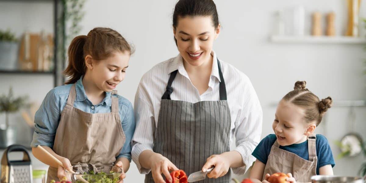 A woman and two young girls, all wearing aprons, prepare food together in a bright kitchen. The woman slices a red pepper while one girl mixes a salad and the other smiles, holding a tomato. Fresh vegetables are on the counter.