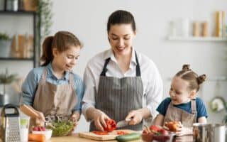 A woman and two young girls, all wearing aprons, prepare food together in a bright kitchen. The woman slices a red pepper while one girl mixes a salad and the other smiles, holding a tomato. Fresh vegetables are on the counter.