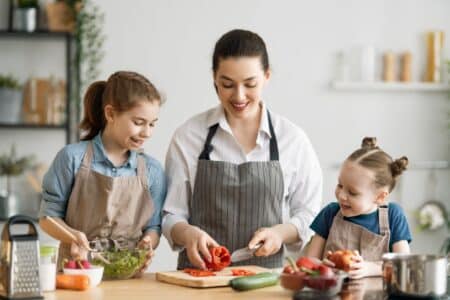 A woman and two young girls, all wearing aprons, prepare food together in a bright kitchen. The woman slices a red pepper while one girl mixes a salad and the other smiles, holding a tomato. Fresh vegetables are on the counter.