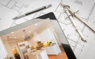 A home floor plan with a pen and compass beside a tablet displaying a photograph of a modern white kitchen with granite worktops, stainless steel appliances, and wooden floors. The setup suggests home design or renovation planning.
