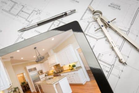 A home floor plan with a pen and compass beside a tablet displaying a photograph of a modern white kitchen with granite worktops, stainless steel appliances, and wooden floors. The setup suggests home design or renovation planning.