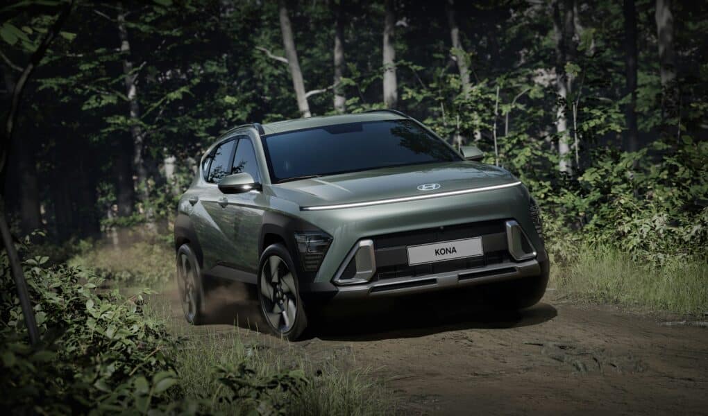 A metallic green Hyundai Kona SUV drives on a dirt track through a lush, green forest. The car kicks up dust and sunlight filters through dense trees in the background, highlighting the Kona’s sleek, modern design and prominent front grille.