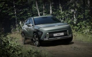 A metallic green Hyundai Kona SUV drives on a dirt track through a lush, green forest. The car kicks up dust and sunlight filters through dense trees in the background, highlighting the Kona’s sleek, modern design and prominent front grille.